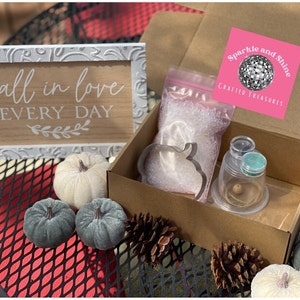 May include: A craft kit with a pumpkin-shaped cookie cutter, clear beads, and a clear glass jar with a teal lid. The kit is packaged in a brown box with a pink sticker that says "Sparkle and Shine Crafted Treasures". The box is sitting on a black metal table with a red surface. There is a wooden sign that says "fall in love every day" and several small pumpkins.