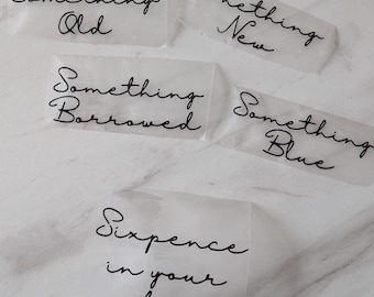 Puede incluir: Cinco etiquetas translúcidas con escritura negra: "Something Old", "Something New", "Something Borrowed", "Something Blue" y "Sixpence in your shoe". Las etiquetas están dispuestas sobre una superficie blanca.