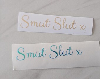 Puede incluir: Dos pegatinas rectangulares con las palabras "Smut Slut x" en escritura cursiva. Una pegatina tiene letras doradas y la otra tiene letras azul-verdes. Las pegatinas están sobre una superficie de color claro.