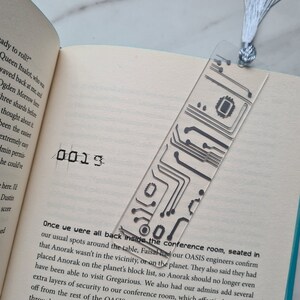 May include: A transparent acrylic bookmark featuring a black circuit board pattern. It has a silver tassel at the top. The bookmark is in an open book. The number "0013" is printed on the page.