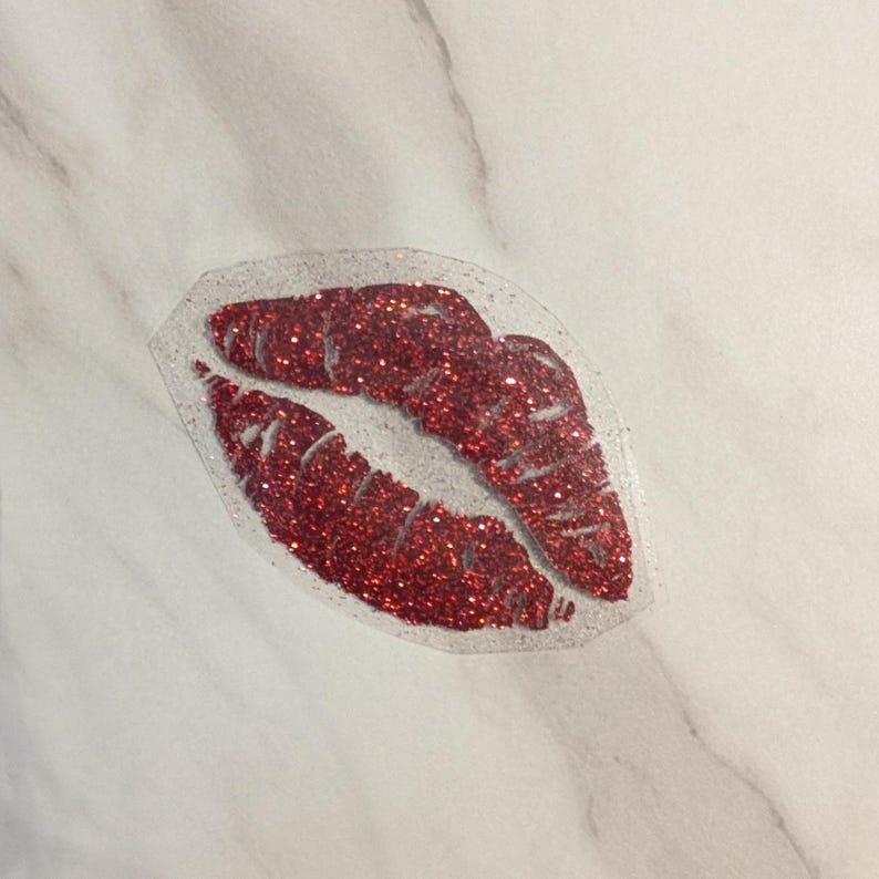Puede incluir: Una pegatina con forma de labios de purpurina roja con un contorno transparente. Los labios est&aacute;n rellenos de purpurina roja brillante, creando un efecto brillante. La pegatina est&aacute; colocada sobre una superficie de m&aacute;rmol blanco.