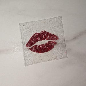 May include: A square, clear sticker featuring a glittery red lip print. The lips have a textured appearance, and the glitter provides a sparkling effect. The sticker is set against a white background.