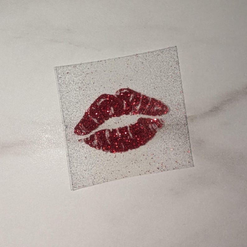 Puede incluir: Una pegatina cuadrada y transparente con un fondo brillante. La pegatina presenta una huella de labios roja con purpurina. Los labios est&aacute;n detallados con l&iacute;neas y rellenos de purpurina roja, creando un efecto brillante.