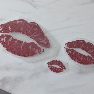 May include: Three red glitter lip-shaped stickers in different sizes. The lips are a deep red colour and have a sparkly texture. The stickers are on a white surface.