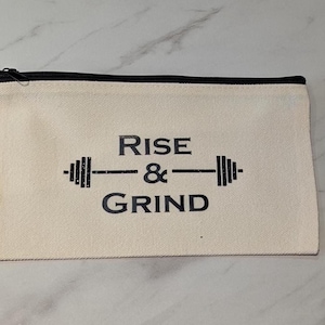 May include: A cream canvas pouch with a black zip, featuring the words "RISE & GRIND" and a barbell graphic in black. The rectangular pouch is likely made from a durable material, suitable for various uses.