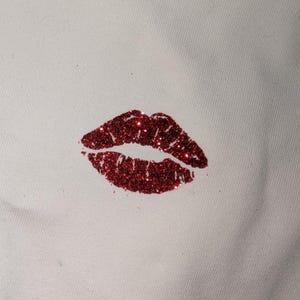 Puede incluir: Primer plano de una tela blanca con un estampado de labios rojos con purpurina. Los labios son de color rojo intenso y est&aacute;n cubiertos de purpurina brillante. El estampado est&aacute; situado en la esquina inferior derecha de la tela.