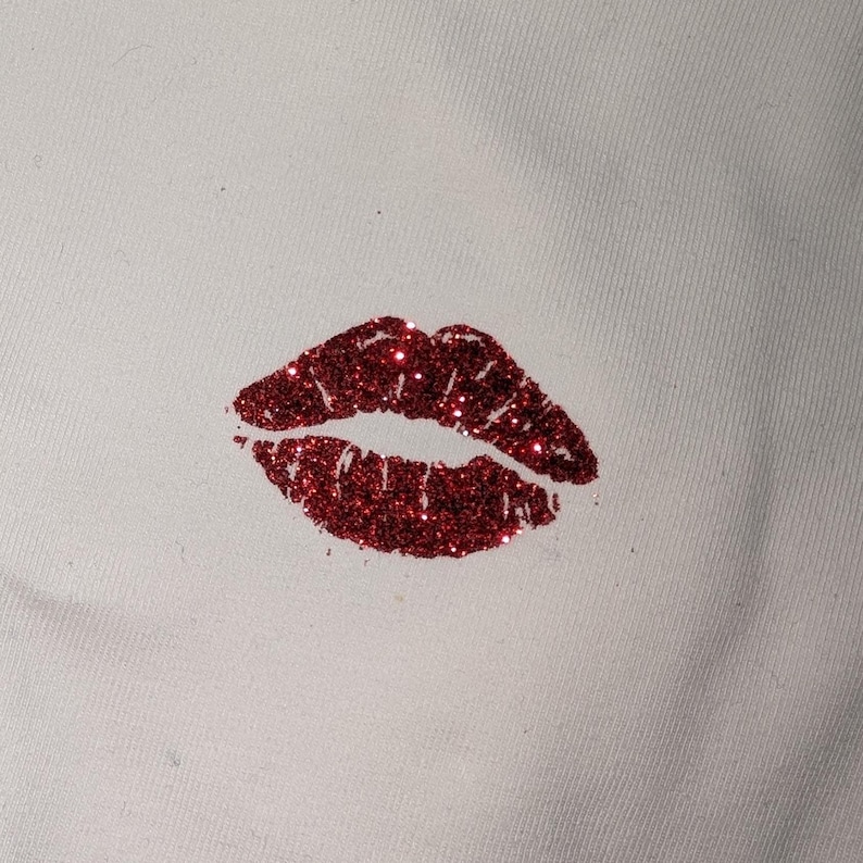 Puede incluir: Primer plano de una tela blanca con un estampado de labios rojos con purpurina. Los labios son de color rojo intenso y est&aacute;n cubiertos de purpurina brillante. El estampado est&aacute; situado en la esquina inferior derecha de la tela.