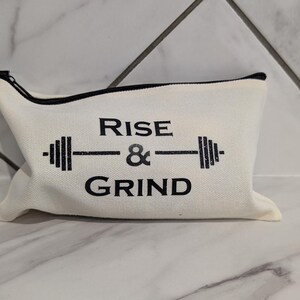 May include: Off-white canvas zippered pouch with black trim. The pouch features the text "RISE & GRIND" in black, with a barbell graphic. The bag is designed for gym or fitness enthusiasts.