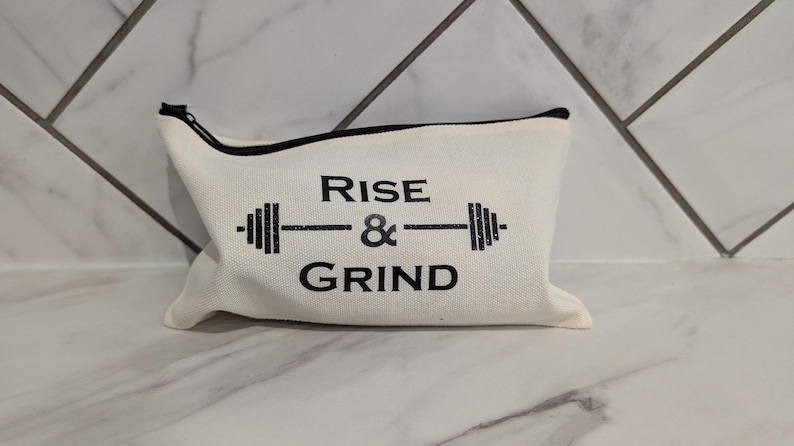 May include: Off-white canvas zippered pouch with black trim. The pouch features the text "RISE & GRIND" in black, with a barbell graphic. The bag is designed for gym or fitness enthusiasts.