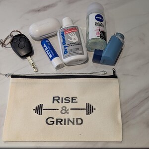May include: An assortment of everyday carry items, including a car key, lip balm, hand lotion, deodorant, a small inhaler, and a white wireless earbud case. A canvas pouch with the text "Rise & Grind" is also present.