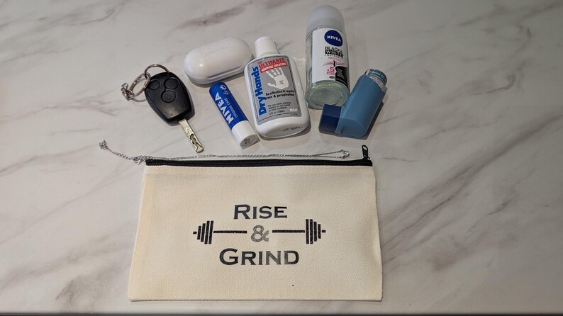 May include: An assortment of everyday carry items, including a car key, lip balm, hand lotion, deodorant, a small inhaler, and a white wireless earbud case. A canvas pouch with the text "Rise & Grind" is also present.