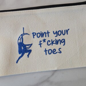 May include: Cream-colored canvas pencil case with a black zipper. The case features a blue illustration of a person on a hoop and the text "point your f*cking toes" in blue lettering. The design is centered on the case.