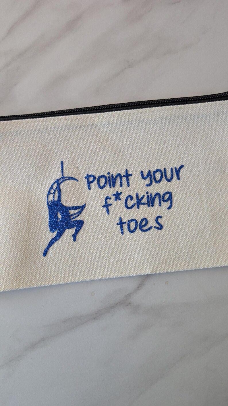 May include: Cream-colored canvas pencil case with a black zipper. The case features a blue illustration of a person on a hoop and the text "point your f*cking toes" in blue lettering. The design is centered on the case.
