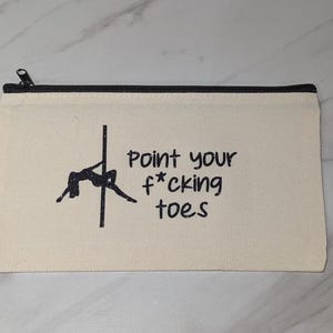 May include: Cream-colored canvas zipper pouch with a black zipper and trim. The pouch features a black silhouette of a person pole dancing and the text "Point your f*cking toes" in black.