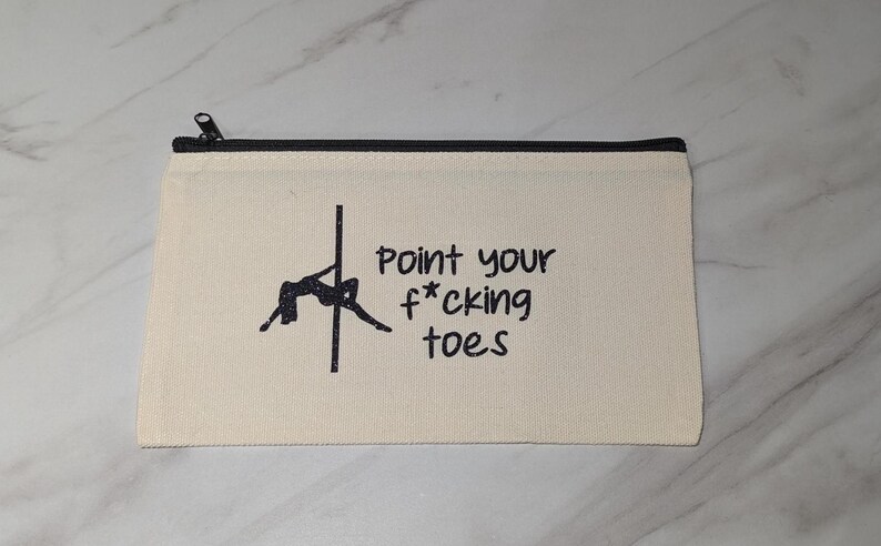 May include: Cream-colored canvas zipper pouch with a black zipper and trim. The pouch features a black silhouette of a person pole dancing and the text "Point your f*cking toes" in black.