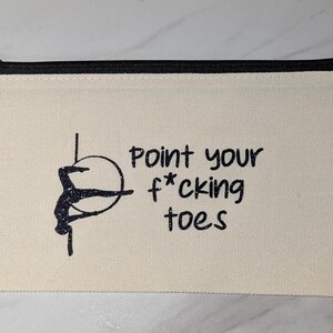 May include: Cream-colored canvas zipper pouch with a black zipper and the text "Point your f*cking toes." A black silhouette of a person on a hoop is on the left side of the text. The pouch is rectangular.