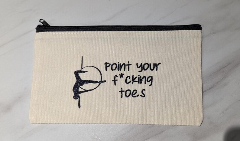 May include: Cream-colored canvas zipper pouch with a black zipper and the text "Point your f*cking toes." A black silhouette of a person on a hoop is on the left side of the text. The pouch is rectangular.