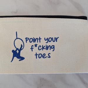 May include: Cream-colored canvas pencil case with a black zipper. The case features a blue graphic of a person on a hoop and the text "Point your f*cking toes" in blue. The case is rectangular.