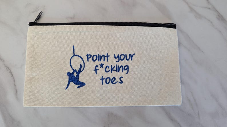 May include: Cream-colored canvas pencil case with a black zipper. The case features a blue graphic of a person on a hoop and the text "Point your f*cking toes" in blue. The case is rectangular.