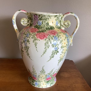 May include: A white ceramic vase with two handles, adorned with hand-painted pink roses, blue and purple flowers, and green foliage. The vase has a wide base and a flared rim, perfect for displaying floral arrangements.
