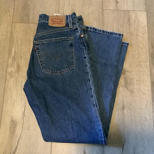 May include: Blue denim jeans with a classic five-pocket design. The jeans feature a brown leather patch on the back waistband and a red tab on the back pocket. The jeans are folded and lying on a wooden surface.
