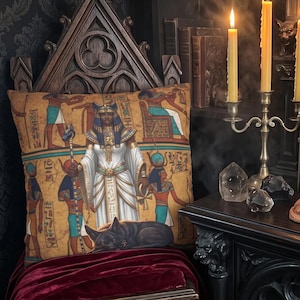 May include: A square throw pillow featuring a vibrant Egyptian-themed print with figures in traditional attire, hieroglyphics, and a black cat. The pillow rests on a dark wooden chair draped with a deep red velvet cloth, surrounded by candles.