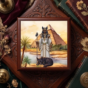 May include: A square wooden box featuring an illustration of an Egyptian deity with a jackal head, wearing a white robe and gold jewelry, holding a staff and an ankh. A black jackal lies at their feet before a pyramid and a river landscape.