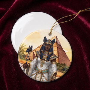 May include: A round ceramic ornament featuring a colorful illustration of an Egyptian deity with a jackal head, wearing a white robe and gold jewelry, holding a staff. The background depicts a large pyramid and palm trees, with a gold hanging cord.