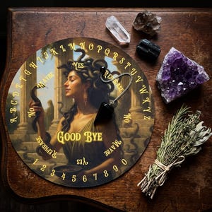 May include: A circular spirit board featuring a Medusa illustration with gold letters A-Z, numbers 0-9, and words Yes, No, Maybe, Rephrase, and Good Bye. A black pointed pendulum on a silver chain rests on the board, accompanied by crystals and herbs.