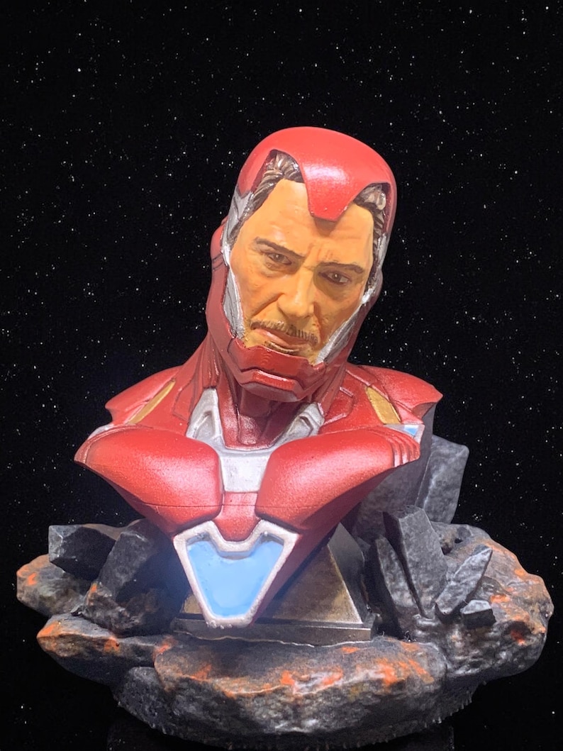 May include: A red and gold Iron Man bust with a detailed face and a blue arc reactor. The bust is on a black and grey rock base.