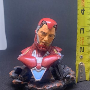 May include: A red, white and blue Iron Man bust figurine with a detailed face and a rocky base. The figurine is approximately 10 cm tall.