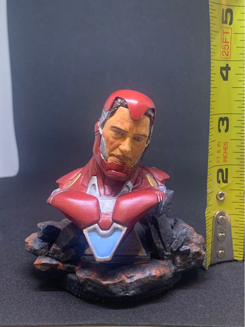 May include: A red, white and blue Iron Man bust figurine with a detailed face and a rocky base. The figurine is approximately 10 cm tall.