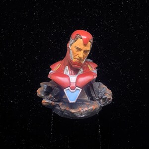 May include: A red and silver bust of Iron Man, a superhero from the Marvel universe, with a distressed rock base. The bust is set against a black background with white stars.