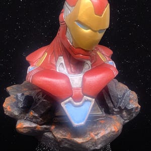 May include: A red and gold Iron Man bust figurine with a blue and silver chest plate. The bust is standing on a rocky base against a black background.