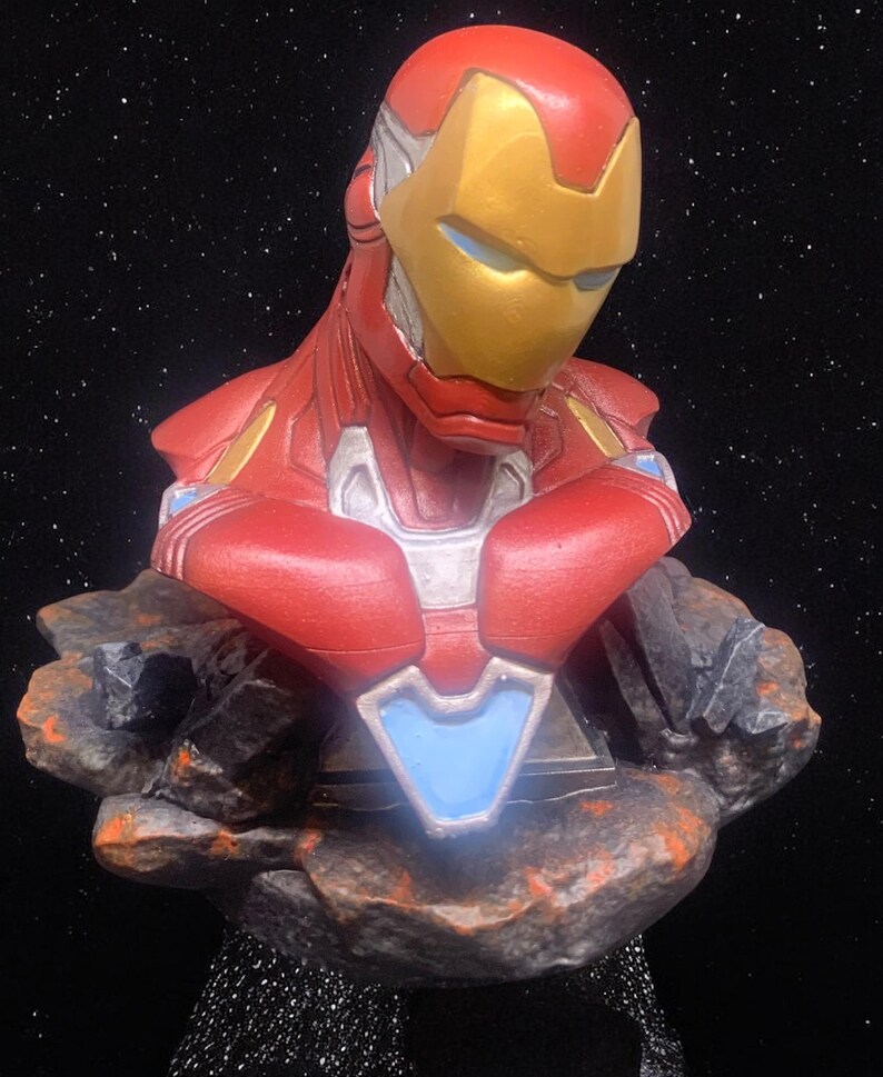 May include: A red and gold Iron Man bust figurine with a blue and silver chest plate. The bust is standing on a rocky base against a black background.