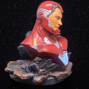 May include: A red and silver bust of Iron Man, a superhero from the Marvel universe, with a detailed face and a textured, rocky base.