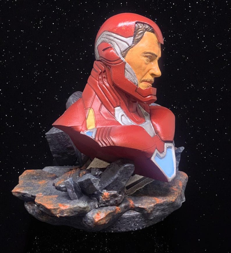May include: A red and silver bust of Iron Man, a superhero from the Marvel universe, with a detailed face and a textured, rocky base.