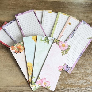 May include: A collection of rectangular paper notepads arranged in a fan shape. Each notepad features horizontal lines, some with checkboxes, and various floral or fruit-themed borders including roses, lilies, and blueberries on white backgrounds.