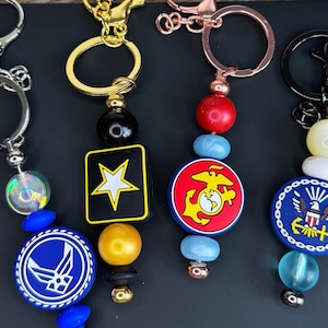 May include: Keychains with military-themed charms. The keychains feature charms with the Air Force, Army, and Marine Corps emblems. Each keychain has a different colored bead and a metal clasp.