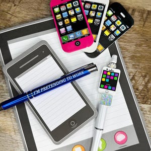 May include: A collection of novelty pens and notepad accessories. The image features a blue pen with the text "I'M PRETENDING TO WORK", along with phone-shaped notepads and pens, including a pink, white, and black phone-shaped pen.