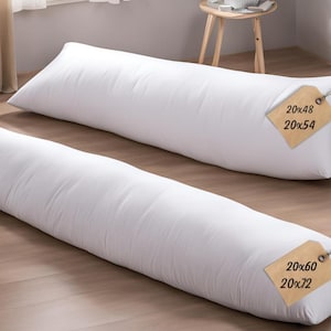 May include: Two white body pillows on a wooden floor. The pillows are labeled with their dimensions: 20x48, 20x54, 20x60, and 20x72 inches. The pillows are made in the USA and are certified by OEKO-TEX Standard 100.