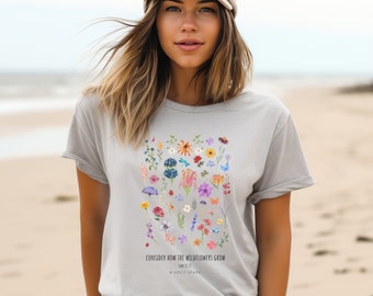 Könnte beinhalten: Ein hellgraues T-Shirt mit einem farbenfrohen Blumendesign mit verschiedenen Wildblumen. Das Shirt enthält den Text "CONSIDER HOW THE WILDFLOWERS GROW" und den Bibelvers Lukas 12,27, mit dem Hashtag #HOLYSPARK.
