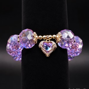 May include: A stretch bracelet featuring large, faceted, translucent purple beads and a central gold-tone heart charm. The heart charm is set with a purple stone and small clear crystals. Gold-tone spacers are placed between the beads.