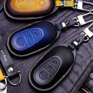 May include: Three leather keychains with different colored and textured leather. The keychains are in the shape of a car key fob. The keychains are on a dark wooden surface.