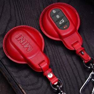 Può includere: Due copri chiavi in ​​pelle rossa per un'auto Mini Cooper. Una copertura ha la parola "MINI" impressa su di essa, mentre l'altra ha quattro pulsanti e un'icona di un'auto. Entrambe le coperture hanno un portachiavi in ​​metallo argentato.