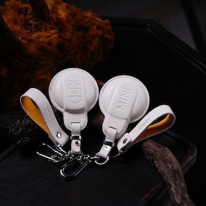 Può includere: Due portachiavi in ​​pelle bianca con anelli metallici. Un portachiavi ha la scritta "MINI" stampata e l'altro ha la scritta "POPPY" stampata.