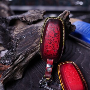 May include: A red and black leather key fob cover with yellow stitching and a silver key ring. The cover has a distressed finish and a matching key fob cover is next to it. The key fob cover is a car accessory.
