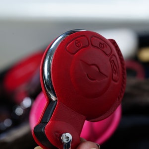 May include: A red leather key fob with a silver trim and a circular button featuring a compass design. The fob has three buttons with symbols for lock, unlock, and boot.