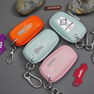 May include: A selection of key fob covers in orange, mint green, and pink, each with a silver-tone keychain. The orange cover has the text "I Mama", the mint green cover has a silhouette design, and the pink cover has the text "Babe".