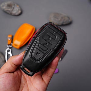 May include: A black leather key fob with red stitching. The fob has four buttons and is designed to fit a car key.